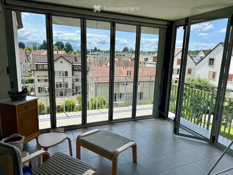 Bright balcony area with large glass doors opening to a view of the neighborhood, featuring two lounge chairs, a small table, and a wooden cabinet with a decorative bowl on top.