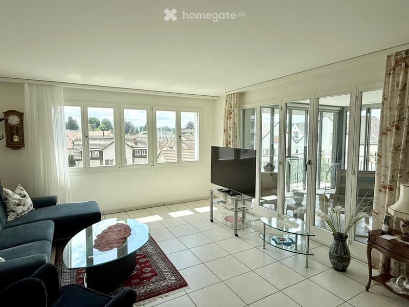 Bright living room with large windows and sliding glass doors leading to a balcony. Features a blue sectional sofa, glass coffee table, flat-screen TV on a glass stand, and decorative items including a vase and a clock on the wall.