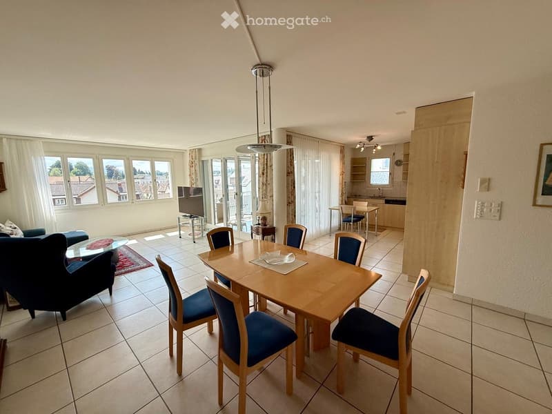 Bright living and dining area with tiled flooring, large windows, and a balcony access. Features a sofa, dining table with chairs, television, and a small kitchen area with a table and chairs.