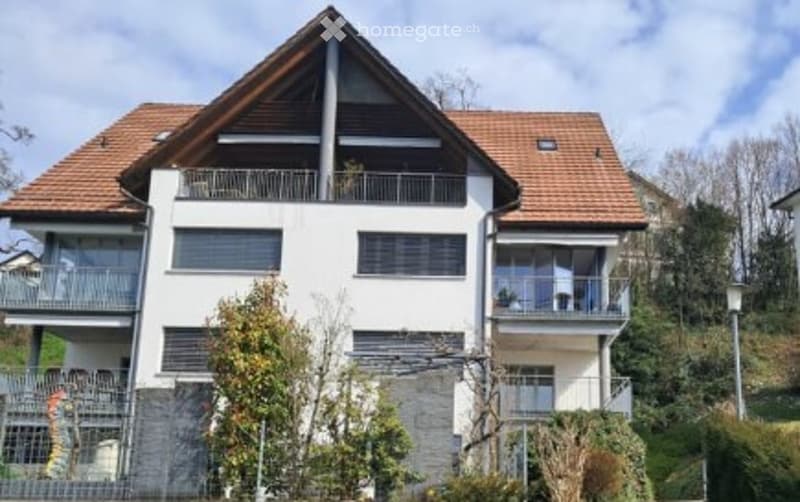Modern two-story house with white walls, dark brown roof, multiple balconies, and surrounding greenery.