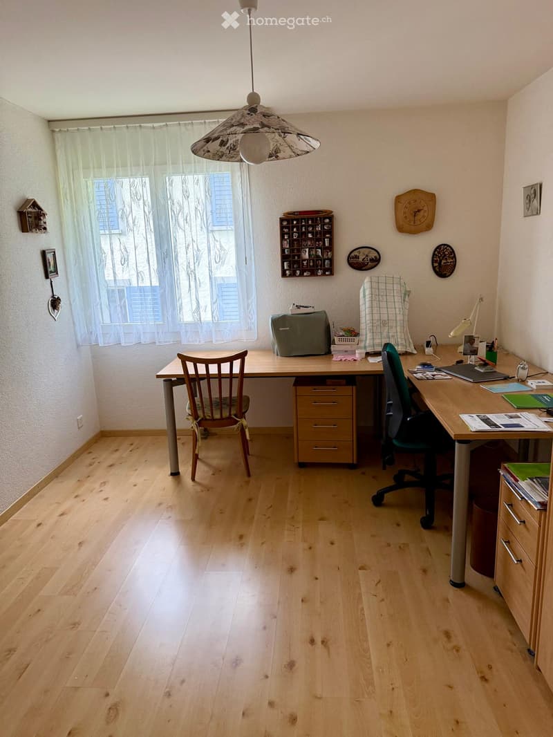 Brightly lit room with wooden flooring, two desks, chairs, a hanging lamp, and various decorative items on the walls.