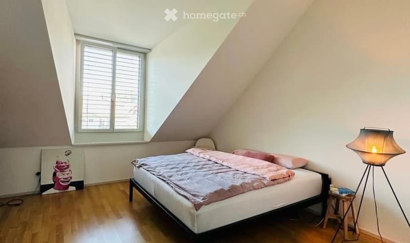 Cozy attic bedroom with wooden flooring, slanted ceilings, large window, bed with pink bedding, nightstand, floor lamp, and artwork on the wall.