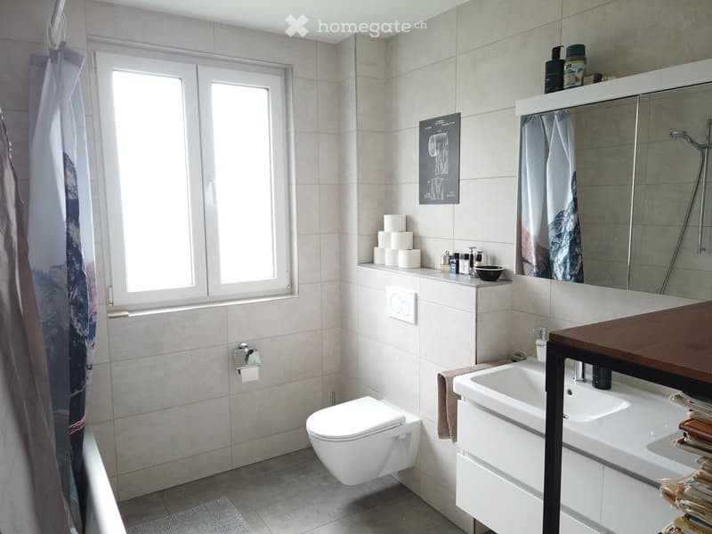 Modern bathroom with white tiles, a toilet, a sink with a wooden countertop, a large mirror, and a shower curtain. Toilet paper rolls and various toiletries are on the shelf.