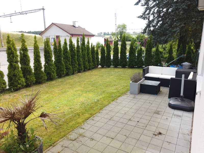 Well-maintained garden with a paved patio area featuring a white sofa, black table, and chairs. The lawn is surrounded by tall, neatly trimmed coniferous trees, and a small house is visible in the background.