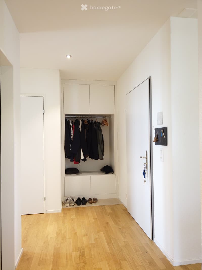 A narrow hallway with light wooden flooring, white walls, and built-in white closet with hanging clothes, shoes, and hats. A door is visible on the right side.
