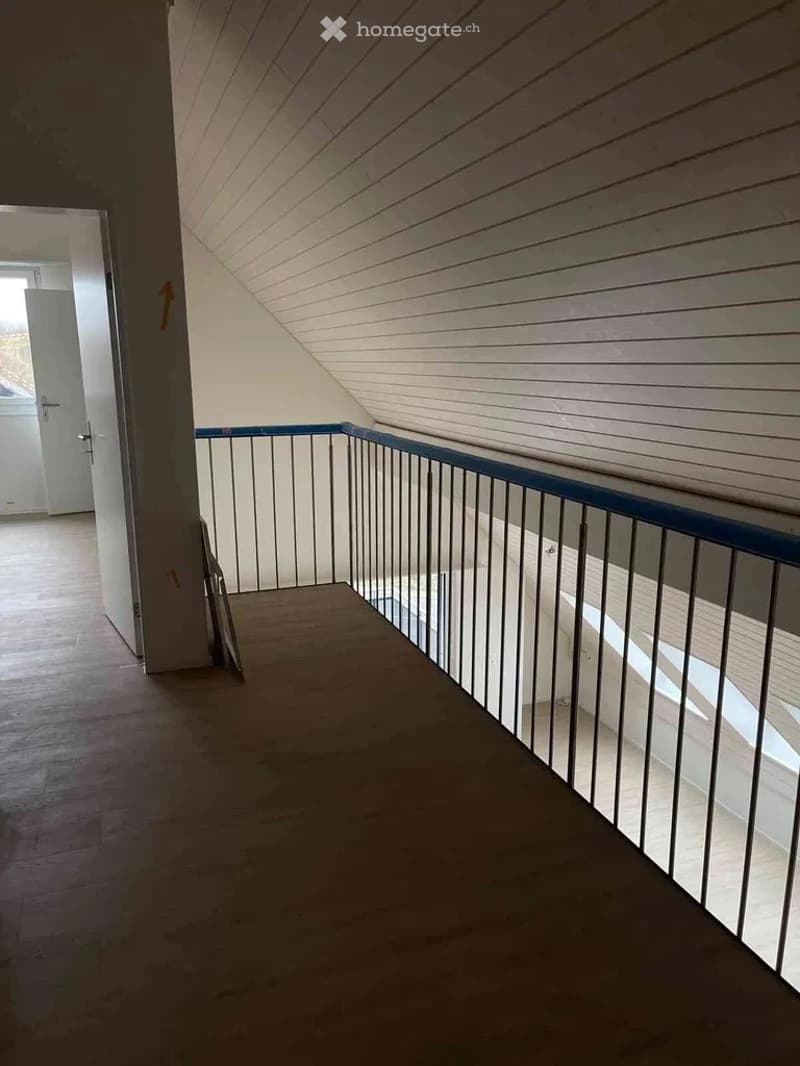 A narrow hallway with wooden flooring and white walls, featuring a metal railing on one side and a door leading to another room at the end.