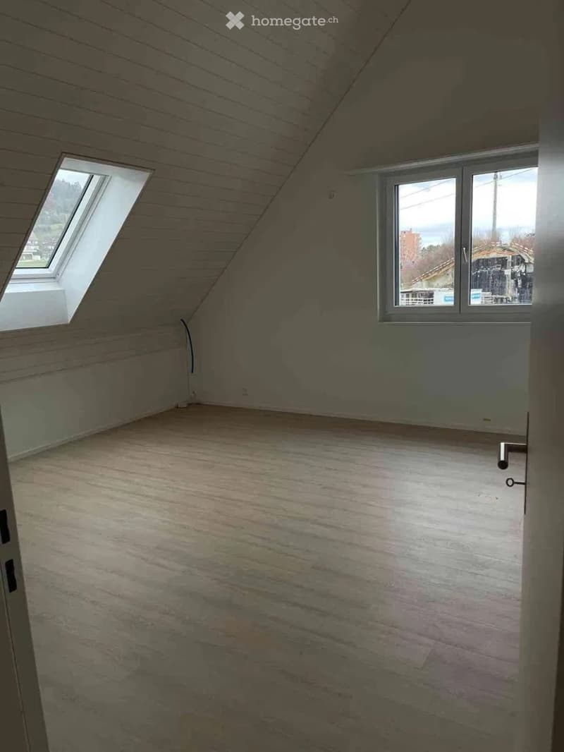 Empty attic room with wooden slanted ceilings, wooden floor, and two windows, one being a skylight.