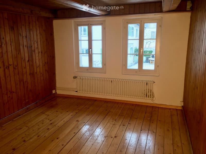 Empty room with wooden flooring, white walls, and two windows with white frames. A radiator is mounted on the wall below the windows.