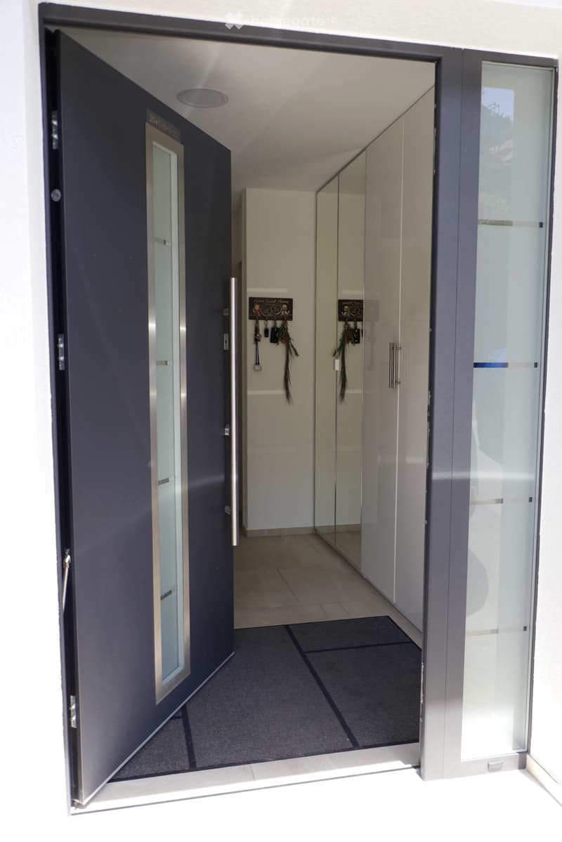 Modern hallway with a dark blue door on the left and a glass-paneled door on the right, leading to another room. The floor is tiled, and the walls are light-colored.