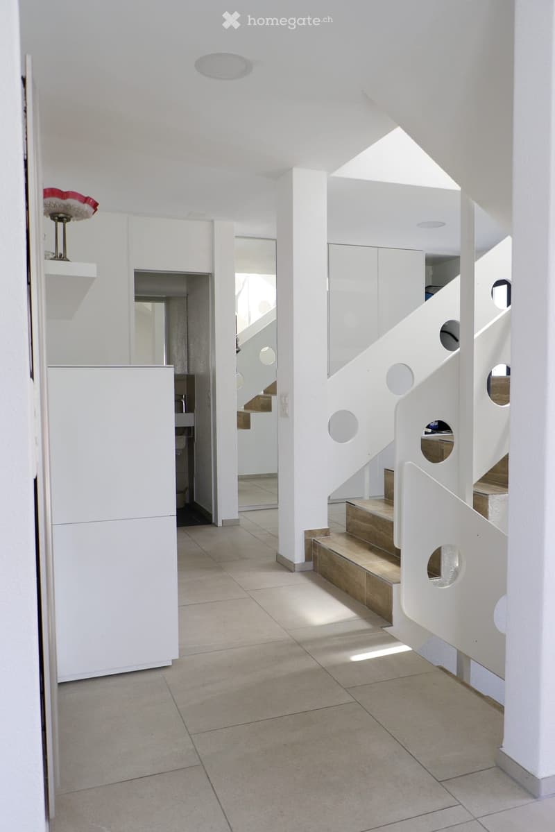 Modern, minimalist hallway with white walls, tiled floor, and a staircase featuring wooden steps and white railings with circular cutouts. A small shelf with a decorative lamp is visible on the left side.