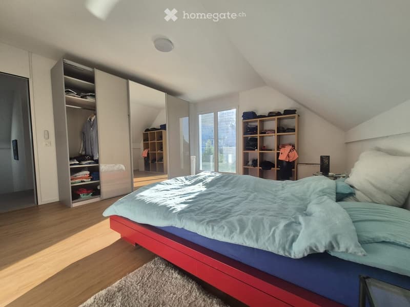 Bright bedroom with wooden flooring, a red-framed bed with blue and light blue bedding, a large mirrored wardrobe, and shelves with folded clothes.