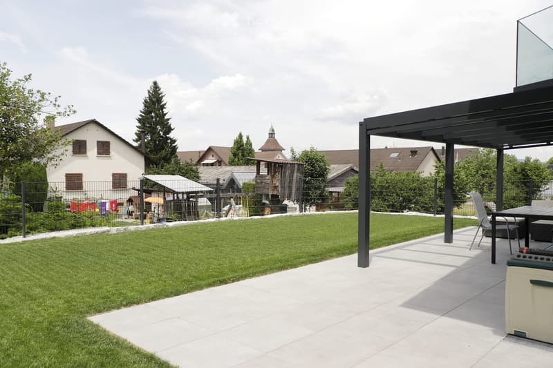 Well-maintained garden with a neatly trimmed lawn, a patio area with a table and chairs, and a modern pergola. The background includes a residential neighborhood with houses and trees.