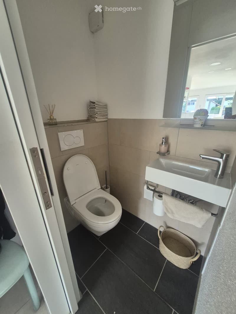 Modern bathroom with a toilet, sink, mirror, and various accessories including a wicker basket, soap dispenser, and towel.