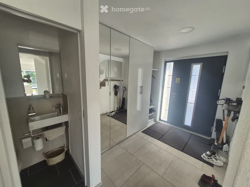 Modern hallway with tiled flooring, large mirror, and adjacent bathroom area visible. Features a sink, mirror, and cleaning supplies in the background.