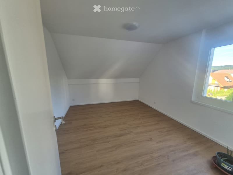 Empty room with wooden flooring, white walls, and a large window offering a view of the outside. The ceiling has a slight slope typical of an attic space.