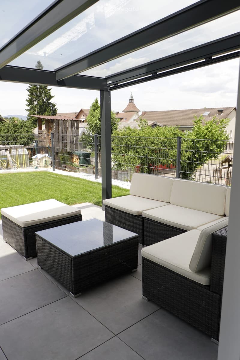 Modern balcony with a wicker sofa set, white cushions, and a glass coffee table, overlooking a fenced garden and houses.
