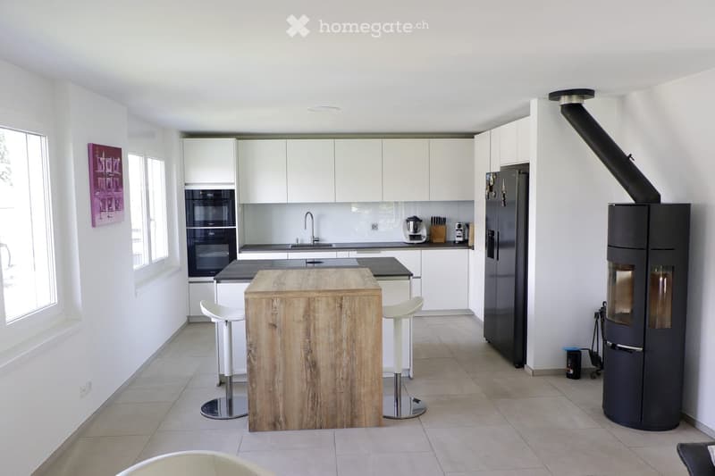 Modern kitchen with white cabinets, a wooden island, two white stools, a black refrigerator, and a built-in oven. A sleek black fireplace is visible on the right side.