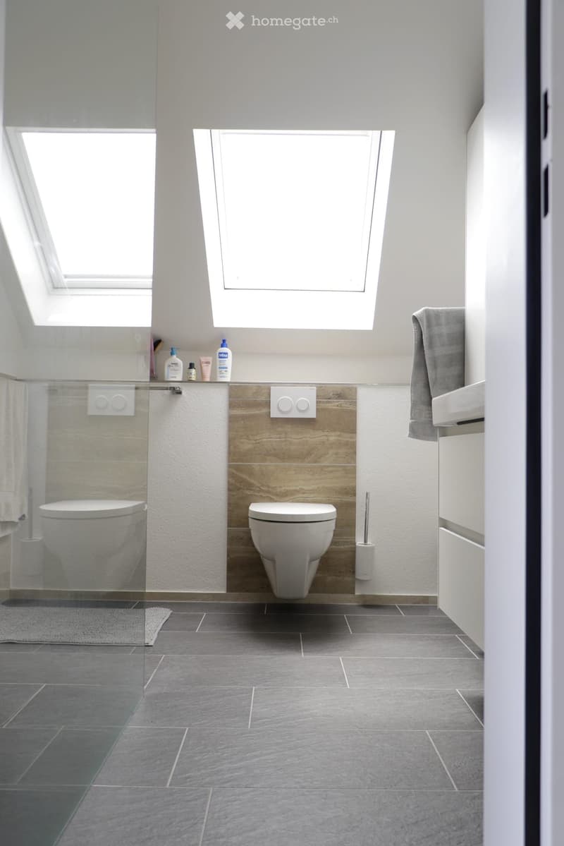 Modern bathroom with two toilets, a sink, a towel, a toilet brush, a shower, and several bottles on the counter. The room features grey tiles and a wooden accent wall.