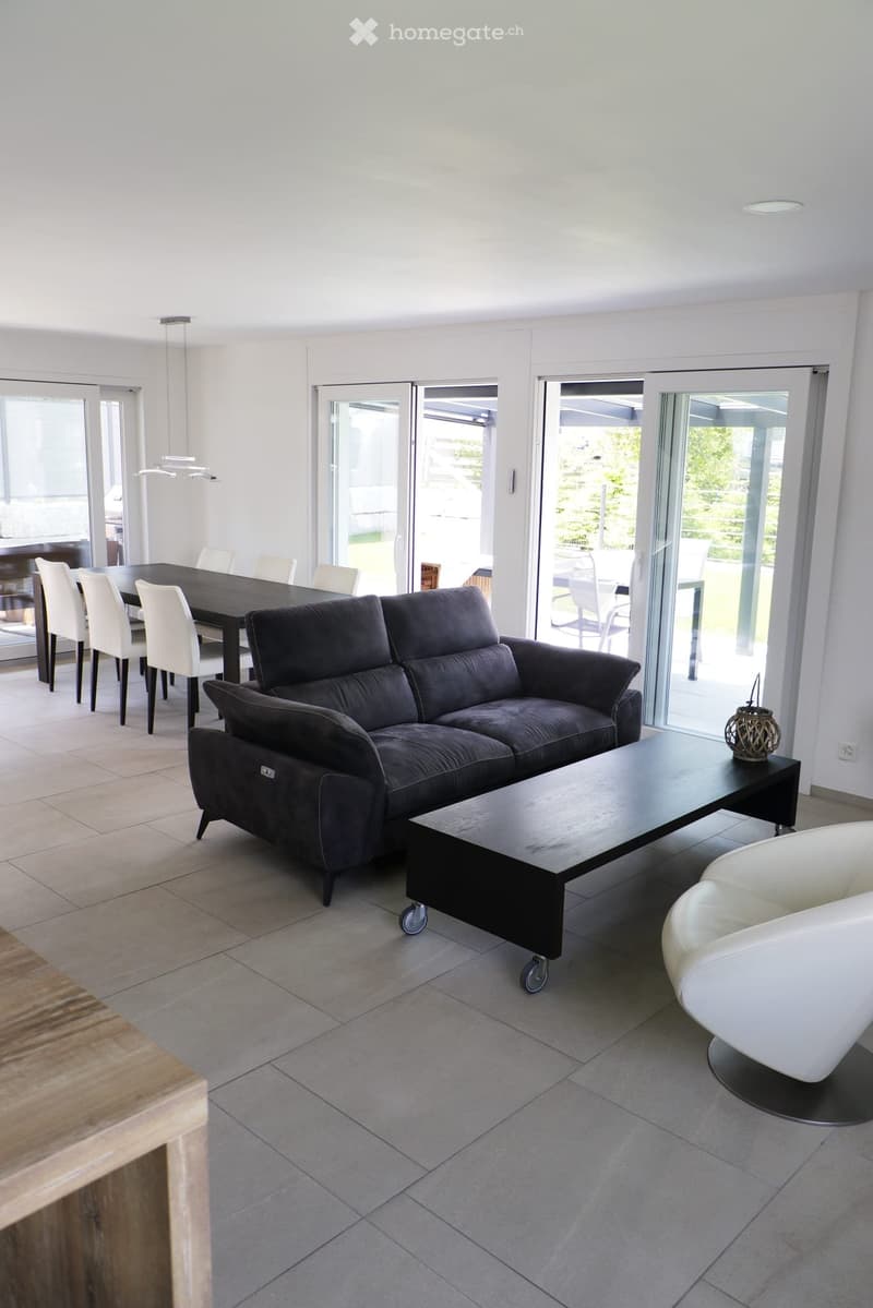 Modern living room with a dark grey sofa, coffee table, white chairs, and a dining table with chairs, large windows and sliding doors leading to a patio.