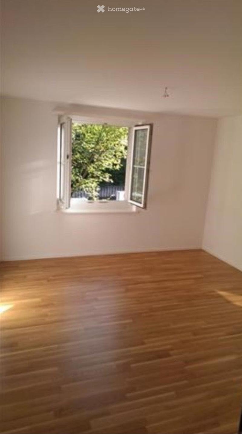 Empty room with wooden flooring and a large window showing greenery outside.