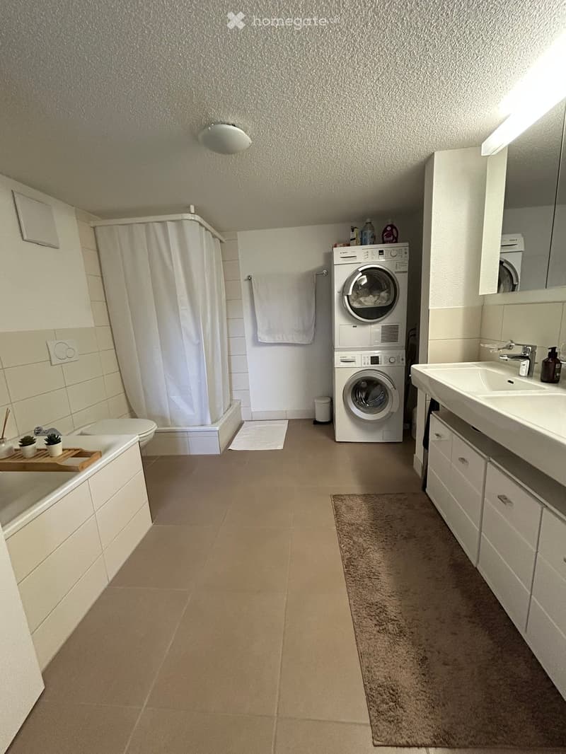Modern bathroom with a bathtub, toilet, sink, washing machine, and a shower area with a curtain. Includes a mirror, towel, and a small rug.