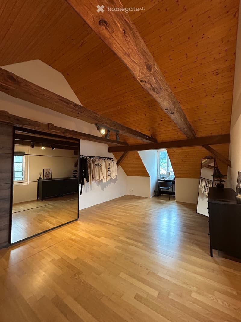 Spacious attic room with wooden beams, wooden flooring, large windows, and visible furniture including a chair, mirror, and cabinet.
