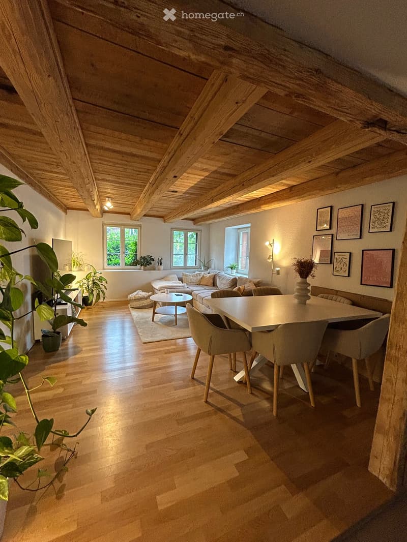 Open-concept living and dining area with exposed wooden beams, hardwood flooring, large windows, sofa, coffee table, dining table, and chairs.