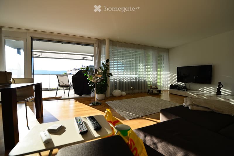 Bright living room with wooden flooring, large windows, and a balcony overlooking the sea. Features a sofa, coffee table, TV, and a fan.