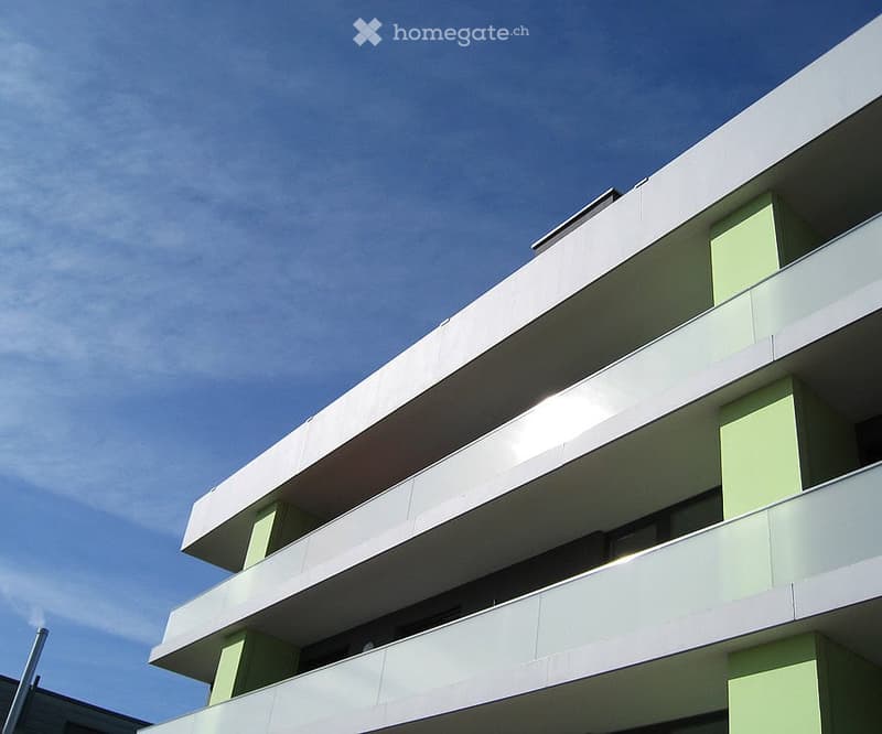 Modern multi-story building with white and green balconies and railings under a clear blue sky.