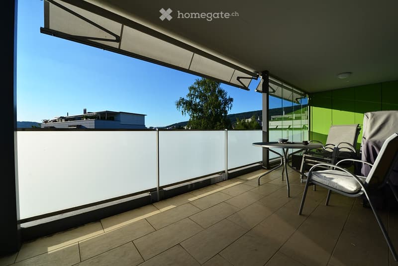 Balcony with tiled floor, white railing, and outdoor furniture including a table and chairs, offering a view of the surrounding area.