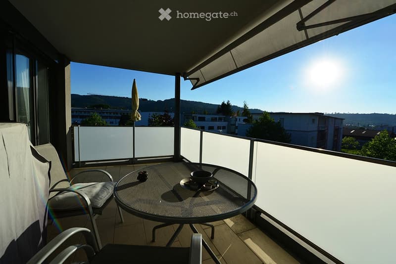 Balcony with a round metal table, two chairs, and an umbrella, overlooking a cityscape with buildings and trees.