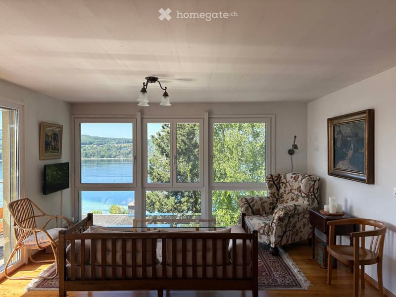 Bright living room with large windows overlooking a lake, featuring a sofa, wicker chair, glass coffee table, armchair, wooden side table, and wall-mounted TV.