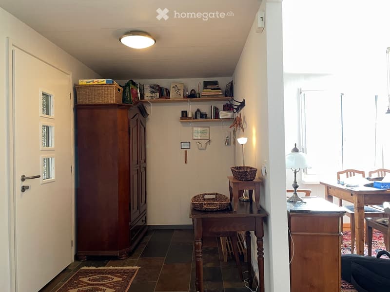 A furnished room with a large wooden wardrobe, a small wooden table with a lamp, a shelf with books and decorative items, and a cozy seating area with a table and chairs.