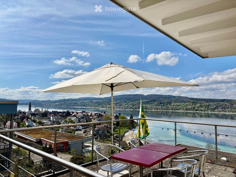 Balcony with a scenic view of a lake and town, featuring a table, chairs, and an umbrella.