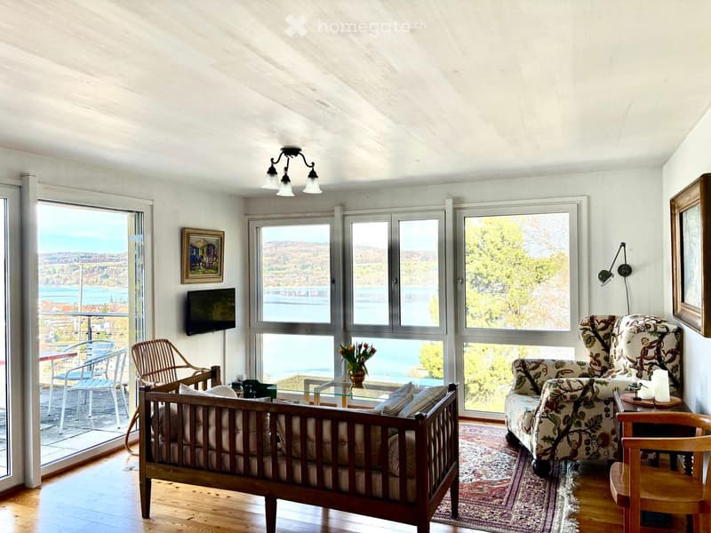 Bright living room with large windows offering a scenic view, featuring a wooden sofa, armchair, coffee table, and a television mounted on the wall.