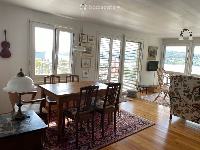 Bright dining area with wooden table, chairs, lamp, television, and scenic view through large windows and sliding doors.