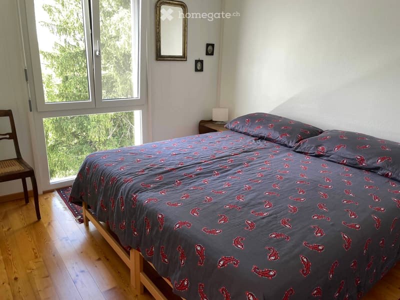 Cozy bedroom with wooden flooring, large windows, a double bed with patterned bedding, a wooden chair, and a nightstand with a lamp.