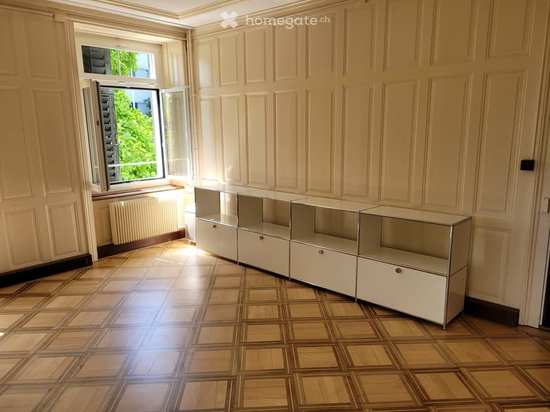 Empty room with cream-colored paneled walls, wooden parquet flooring, and a large window with open shutters. A white radiator is placed beneath the window, and a set of white cabinets with glass doors is positioned against the wall.