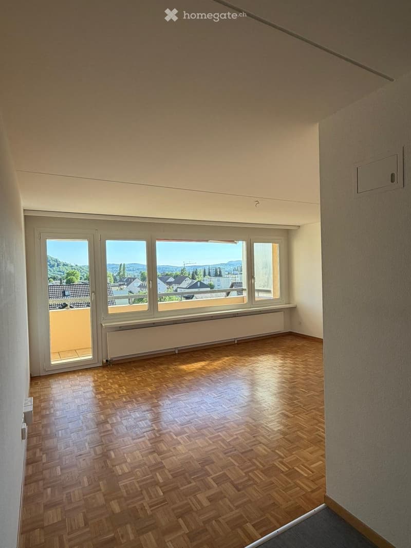 Empty room with wooden parquet flooring, large windows, and a balcony. The room has white walls and a white ceiling.