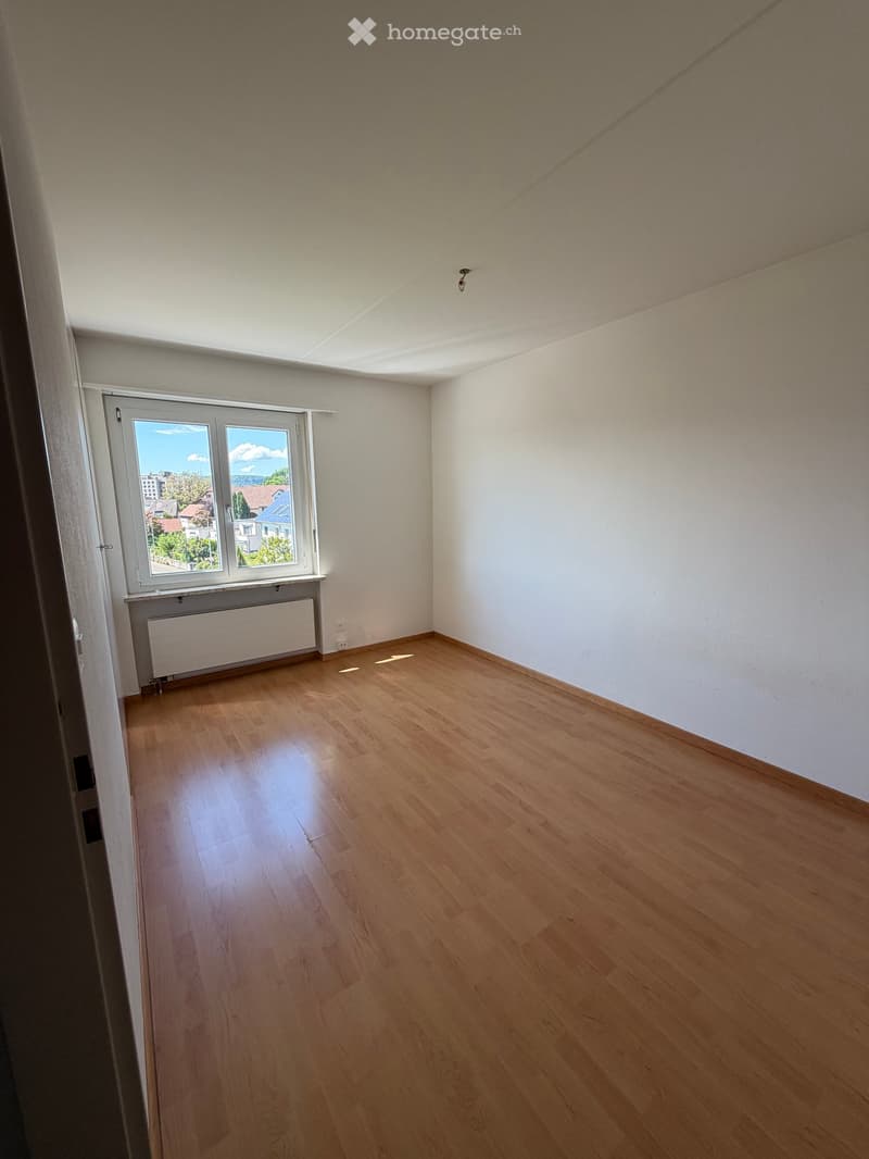 Empty room with wooden flooring, white walls, and a large window offering a view of the outside. A radiator is visible below the window.