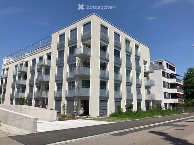 Modern multi-story building with balconies, wide facade, concrete sidewalk, paved street, green lawn, nearby road