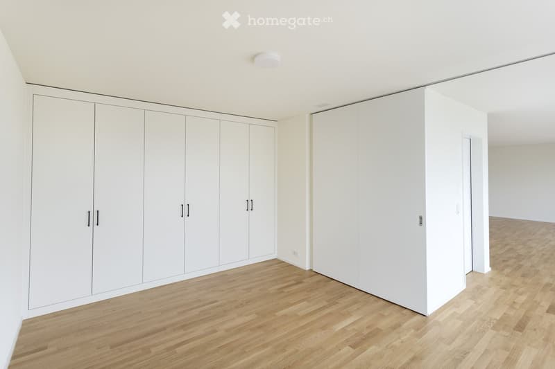 Completely empty room with white walls, light wooden flooring, and built-in white wardrobes.