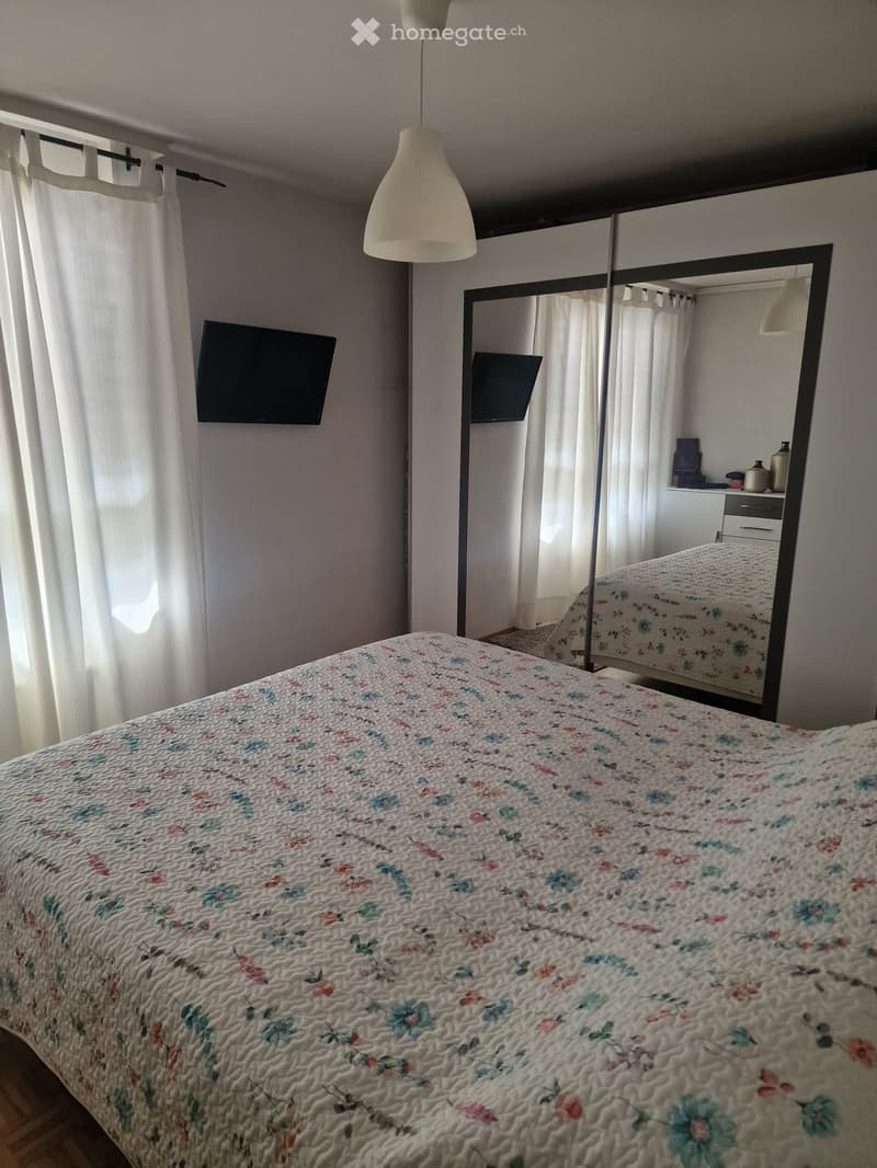A well-lit bedroom with a large bed covered in a floral quilt, a wall-mounted TV, a large mirror reflecting part of the room, and a hanging lamp above the bed.