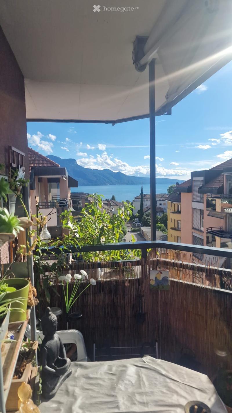 Balcony with a scenic view of a lake and mountains, featuring a table, chair, statue, plants, and an umbrella.