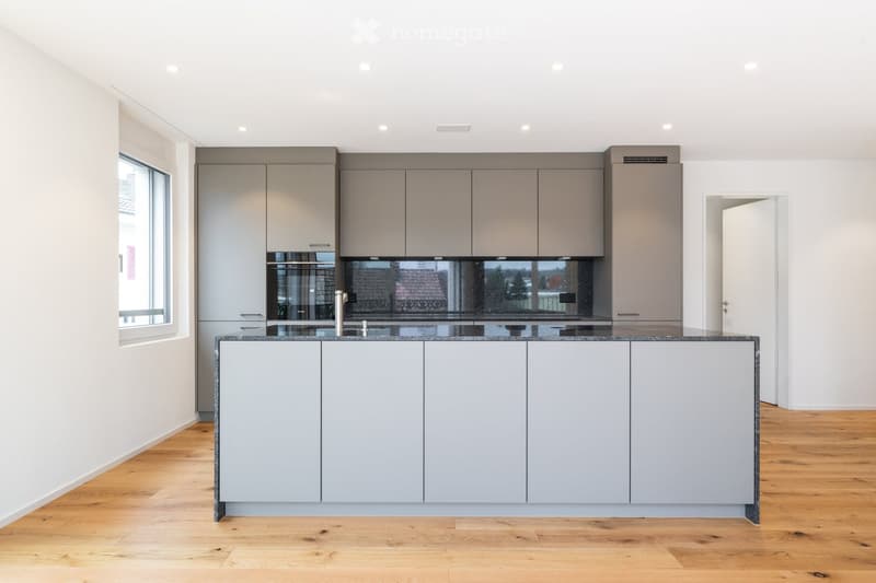 Modern kitchen with grey cabinets, a central island, and a sleek countertop. The space features wooden flooring and large windows allowing natural light to fill the room.
