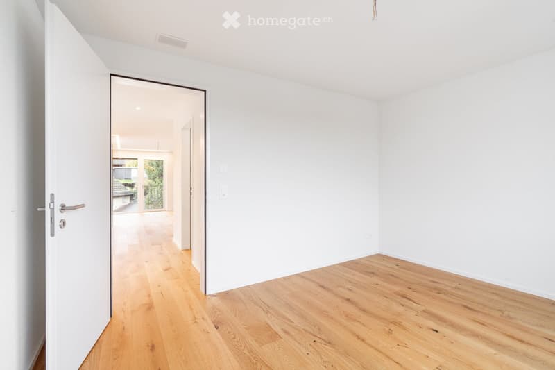 Empty room with white walls and light wooden flooring, featuring an open doorway leading to another space with a view of outdoor greenery.