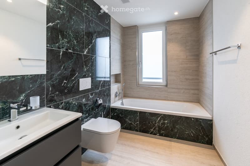 Modern bathroom with dark green marble walls, white fixtures, and a large window. Features a sink, toilet, bathtub, and mirror.