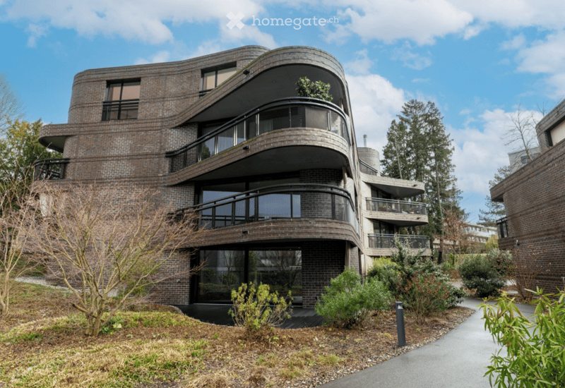 Modern, curved brick building with multiple levels, large windows, and balconies overlooking a landscaped garden area.