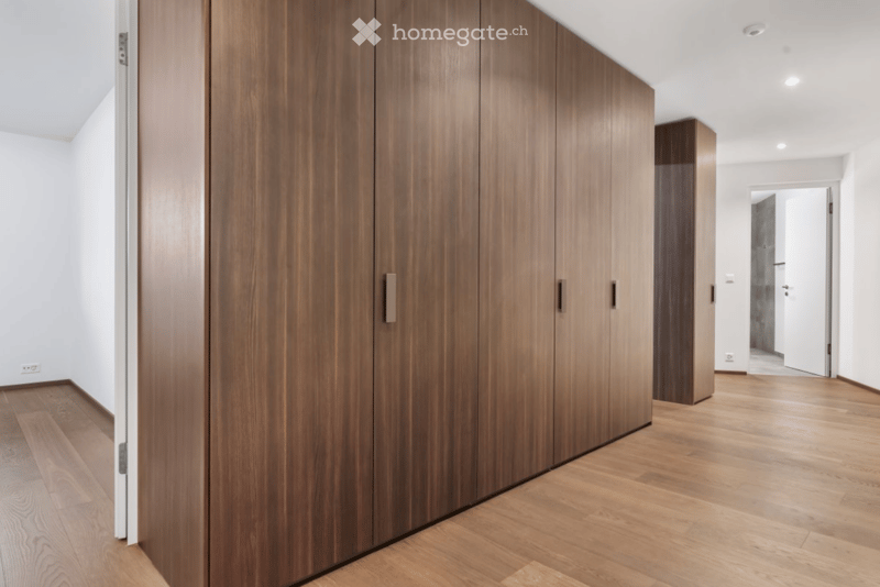 Modern hallway with wooden floor, white walls, and large wooden wardrobe on the left side. The hallway leads to another room with visible white walls and a door.