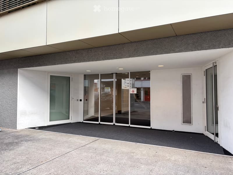 Modern building entrance with glass sliding doors, white walls, and a covered walkway.